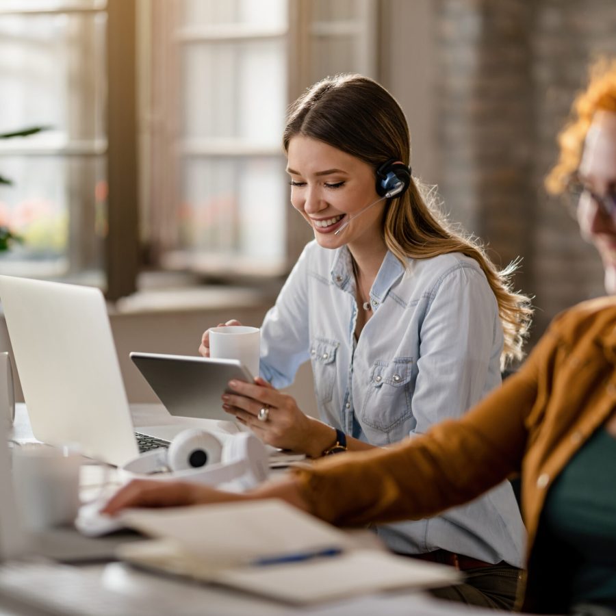 Happy female entrepreneur with headset drinking coffee while surfing the net on touchpad in the office.